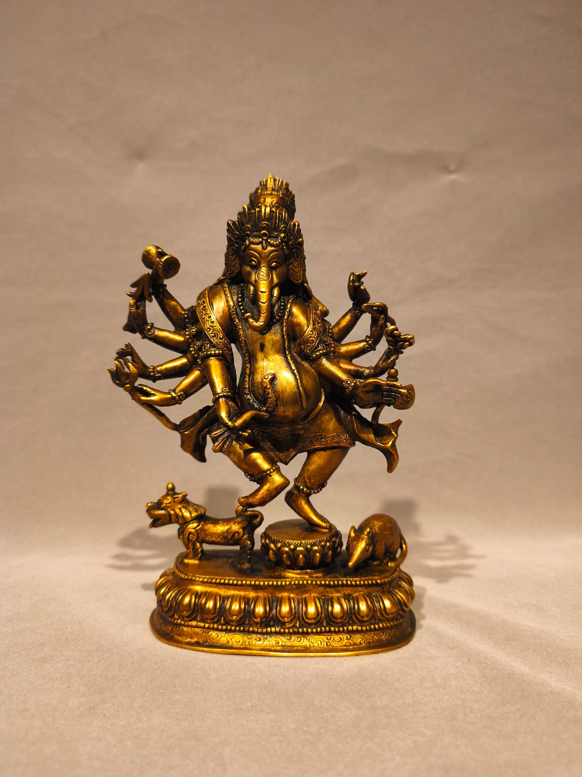 A golden brass statue of Lord Ganesha, the Hindu deity representing wisdom and discipline, standing on a decorated pedestal with a cow and elephant at his base, set against a neutral beige background.