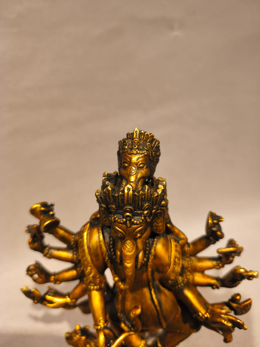 A golden brass statue of a multi-armed Ganesha figure, photographed against a plain light beige background.