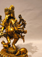 A golden brass statue of Lord Ganesha standing on a decorated pedestal with a small elephant at his side, photographed against a neutral light gray background.