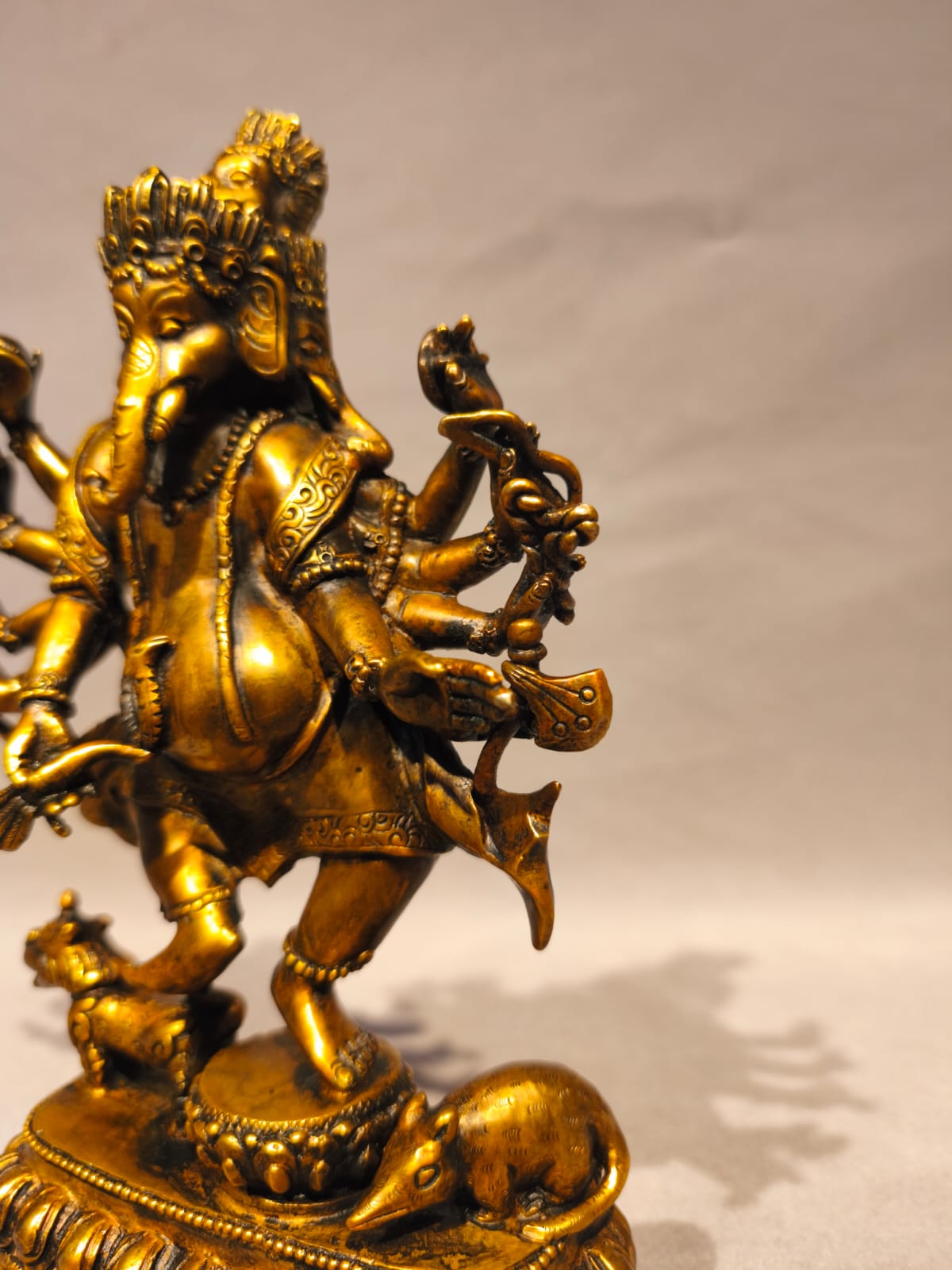 A golden brass statue of Lord Ganesha standing on a decorated pedestal with a small elephant at his side, photographed against a neutral light gray background.