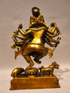 A golden brass statue of a multi-armed dancing deity, shown from the back, mounted on a rectangular base with small animal figures at the bottom, photographed against a neutral light gray background.