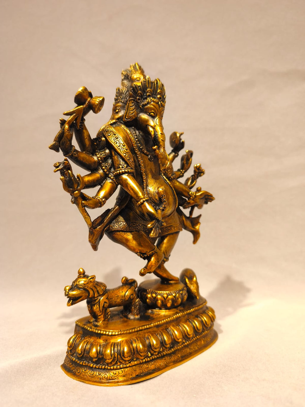 A golden brass statue of Lord Ganesha, the Hindu deity representing wisdom and discipline, standing on a decorated pedestal with a small animal at the front, set against a plain light background.