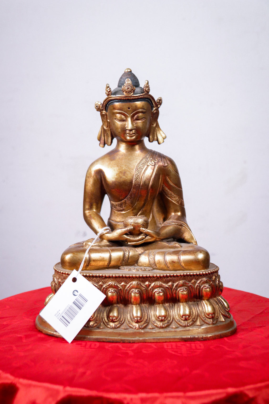 A golden seated Buddha statue on an ornate lotus base, displayed on a red fabric-covered surface against a plain light gray background. A white product tag is attached near the base.