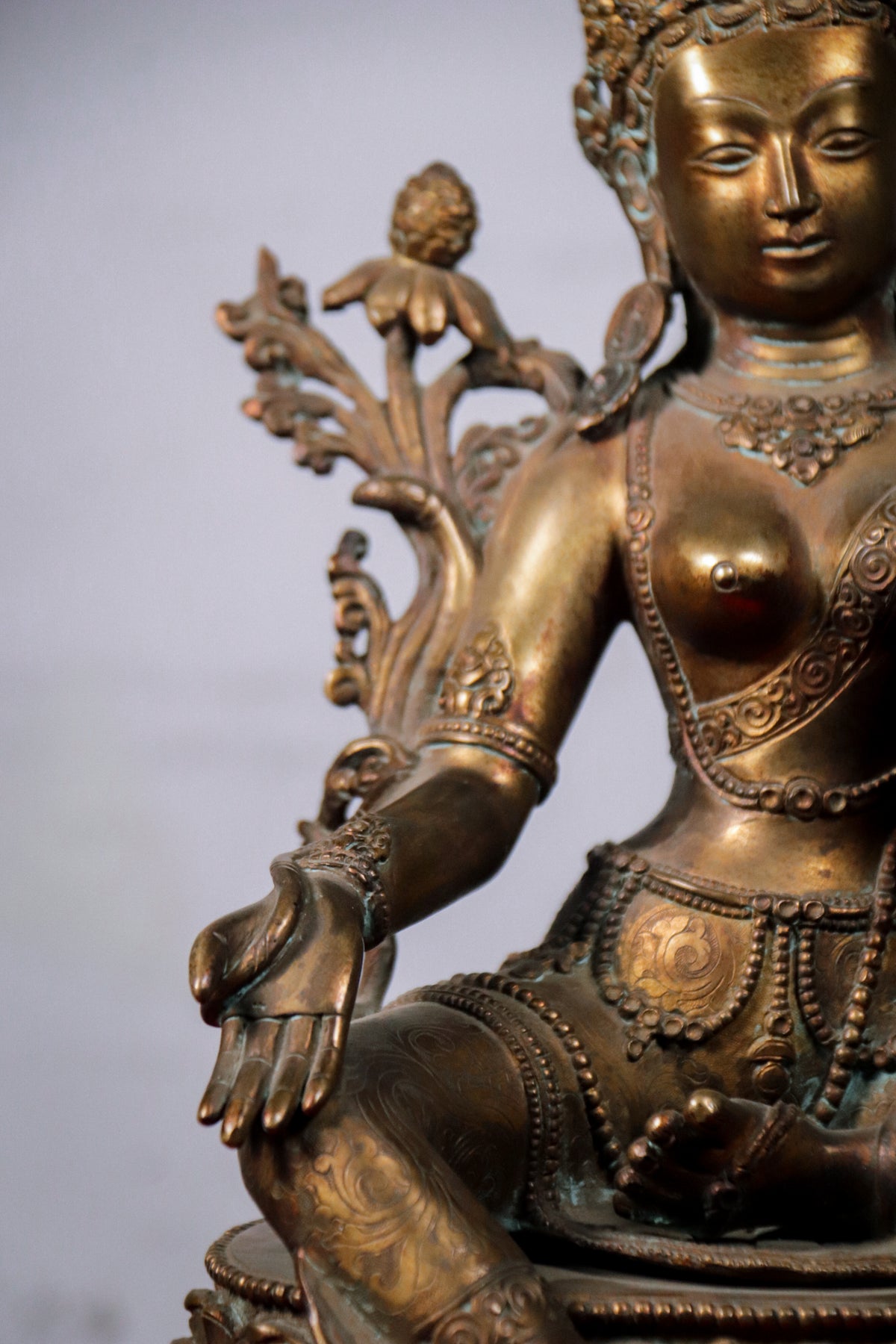 Close-up of an ornate bronze-toned seated statue with detailed sculpted jewelry, floral motifs, and a soft gray background.