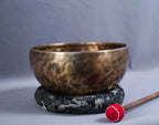 A hammered bronze singing bowl with a carved emblem on the front, resting on a black and gold patterned cushion. A wooden mallet with a red wrapped tip lies nearby on a gray studio background.