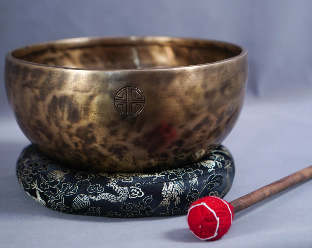 A hammered bronze singing bowl resting on a dark blue patterned cushion, with a wooden mallet featuring a red felt tip placed in front, set against a soft gray background.