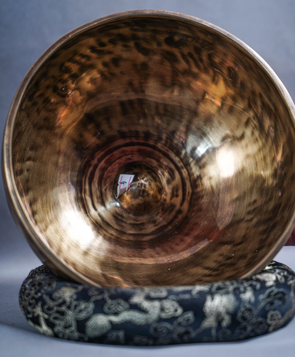 A large metallic singing bowl with a hammered bronze finish, displayed on a dark blue and white patterned fabric stand against a gray background.