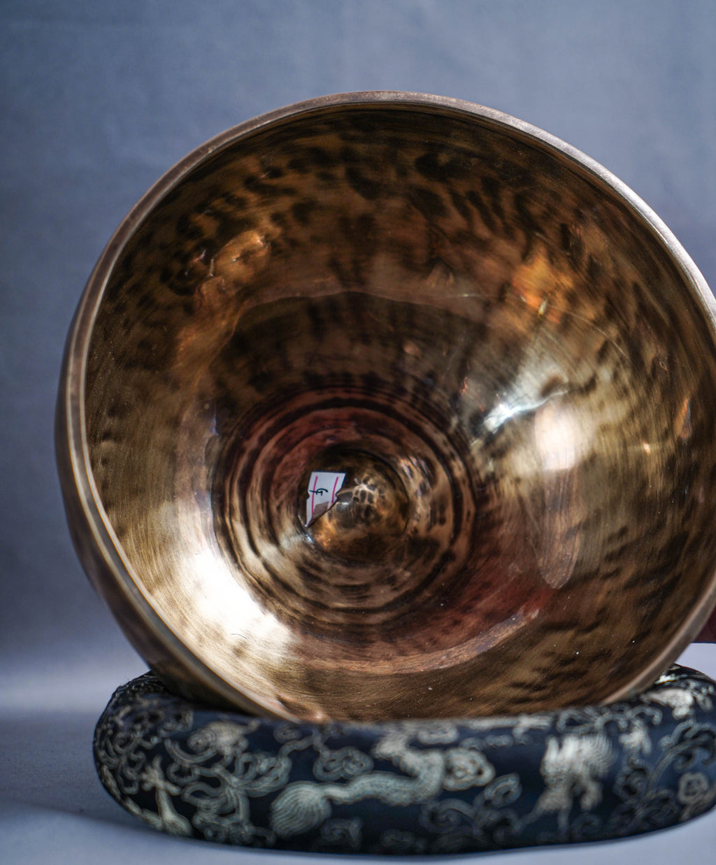 A large hammered bronze singing bowl resting on a dark patterned cushion against a textured gray background.