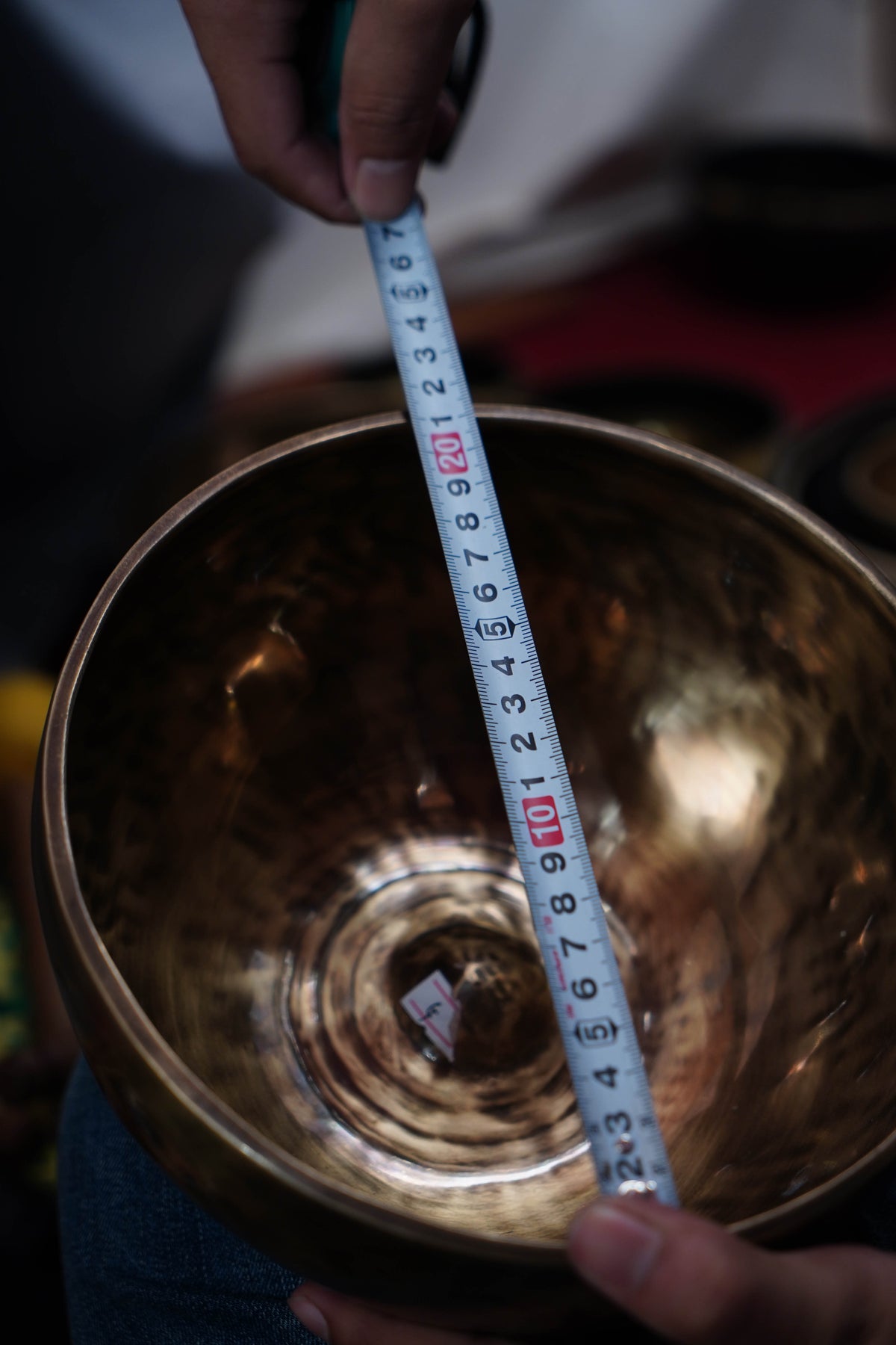 A brass singing bowl being measured with a tape measure held by hands, showing the interior diameter of the bowl.