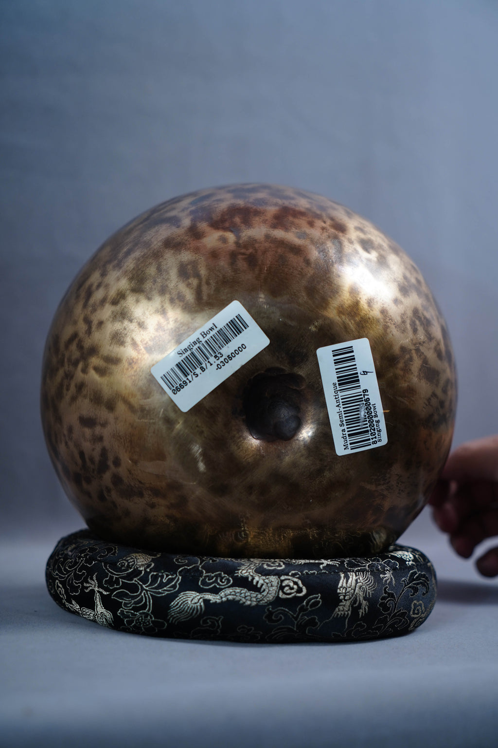 A large, round, hammered metallic gong or temple bell with a mottled bronze and brown finish, resting on a black decorative base with silver dragon motifs. Two white barcode stickers are attached to the front of the gong.