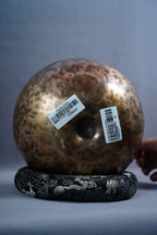 A large, round, hammered metallic gong or temple bell with a mottled bronze and brown finish, resting on a black decorative base with silver dragon motifs. Two white barcode stickers are attached to the front of the gong.
