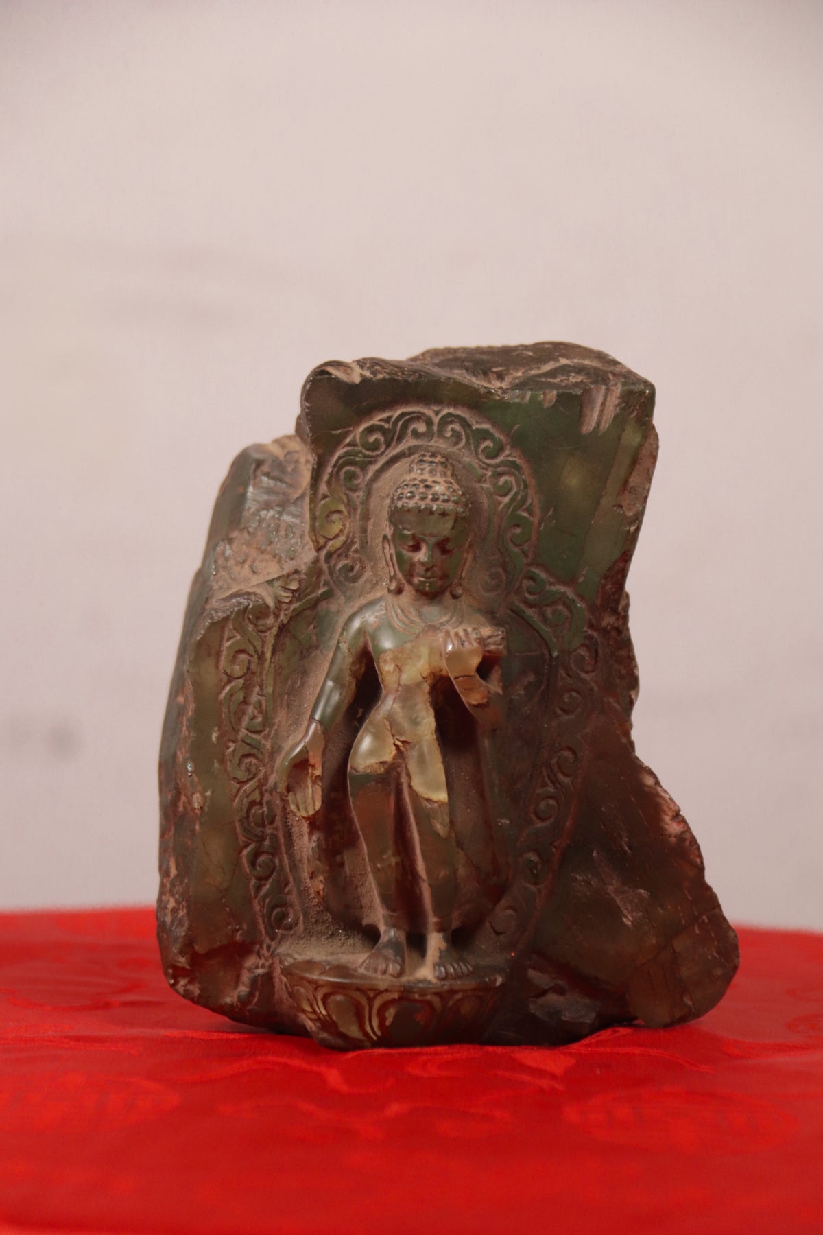 An antique bronze Buddha statue standing on a carved stone slab, displayed on a red surface against a light gray background.